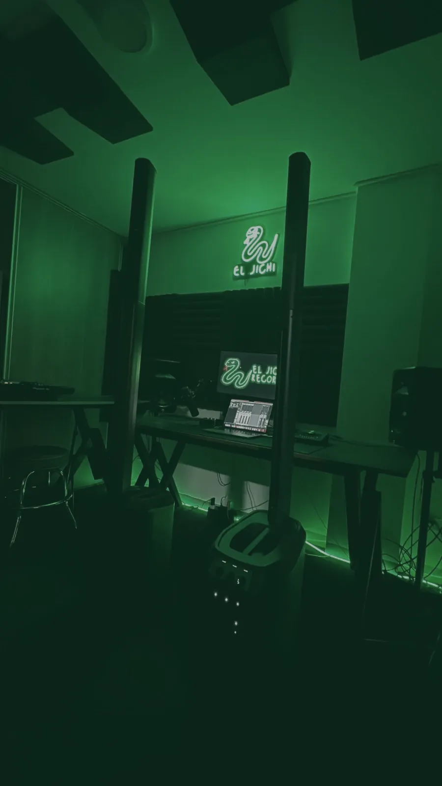 Estudio de El Jichi en luz verde, con los parlantes columna, la mesa de producci&oacute;n y el letrero de ne&oacute;n al fondo